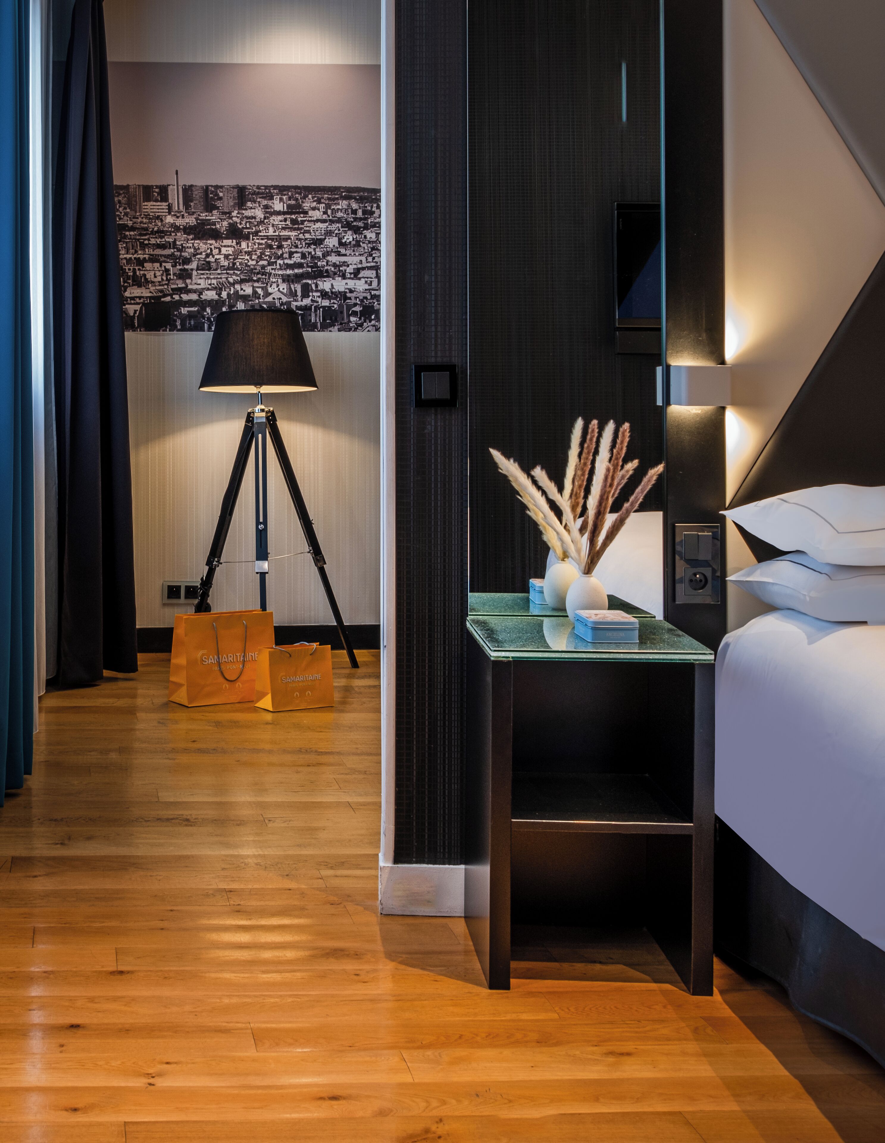 Een hotelkamer met een bed, een nachtkastje met een lamp en decoratieve objecten, een aan de muur bevestigde televisie en uitzicht op een stadslandschap.