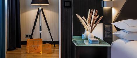 A hotel room with a bed, a nightstand with a lamp and decorative items, a wall-mounted TV, and a cityscape view.
