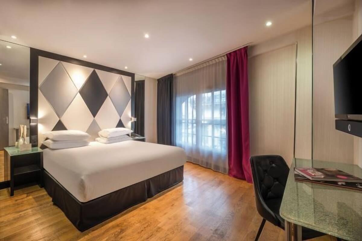 A modern hotel room with a large bed, a glass table, a TV, and a window with curtains.