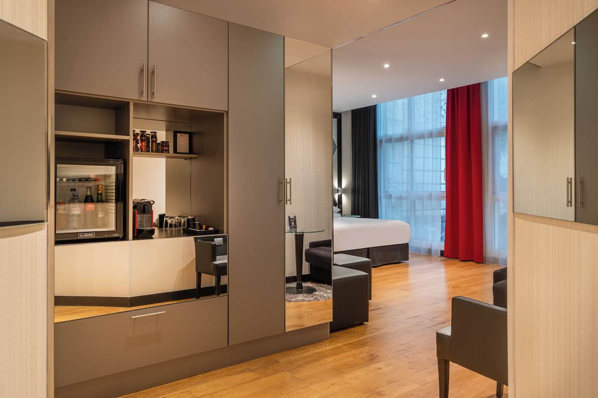 A modern hotel room with a kitchenette, a bed, a seating area, and a large window with curtains.