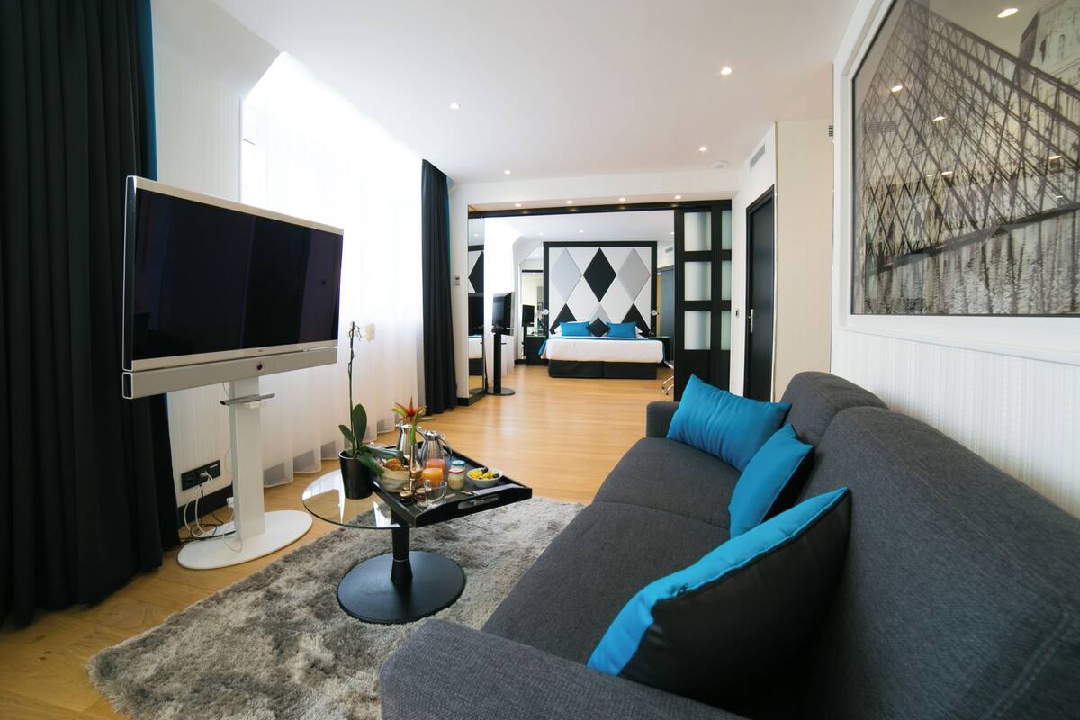 A modern living room with a flat-screen TV, a grey sofa with blue cushions, a coffee table with a tray of items, and a large window with a geometric pattern.