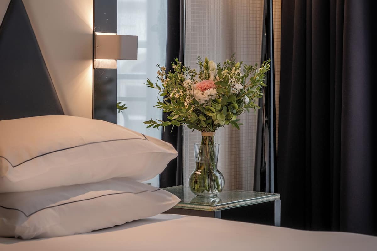 A modern hotel room with a bed, a bedside table with a vase of flowers, a floor lamp, and a large window with curtains.