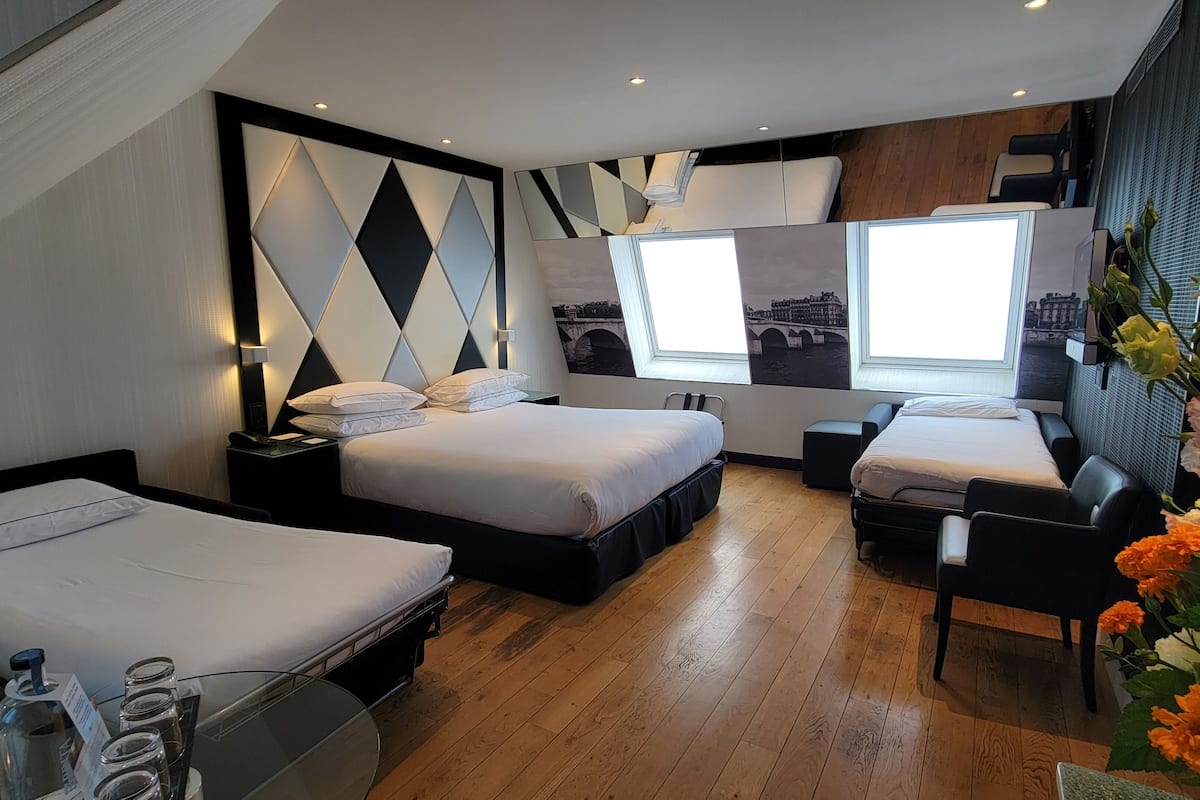 A modern hotel room with two beds, a desk, a chair, a window with a cityscape view, and a decorative wall panel.