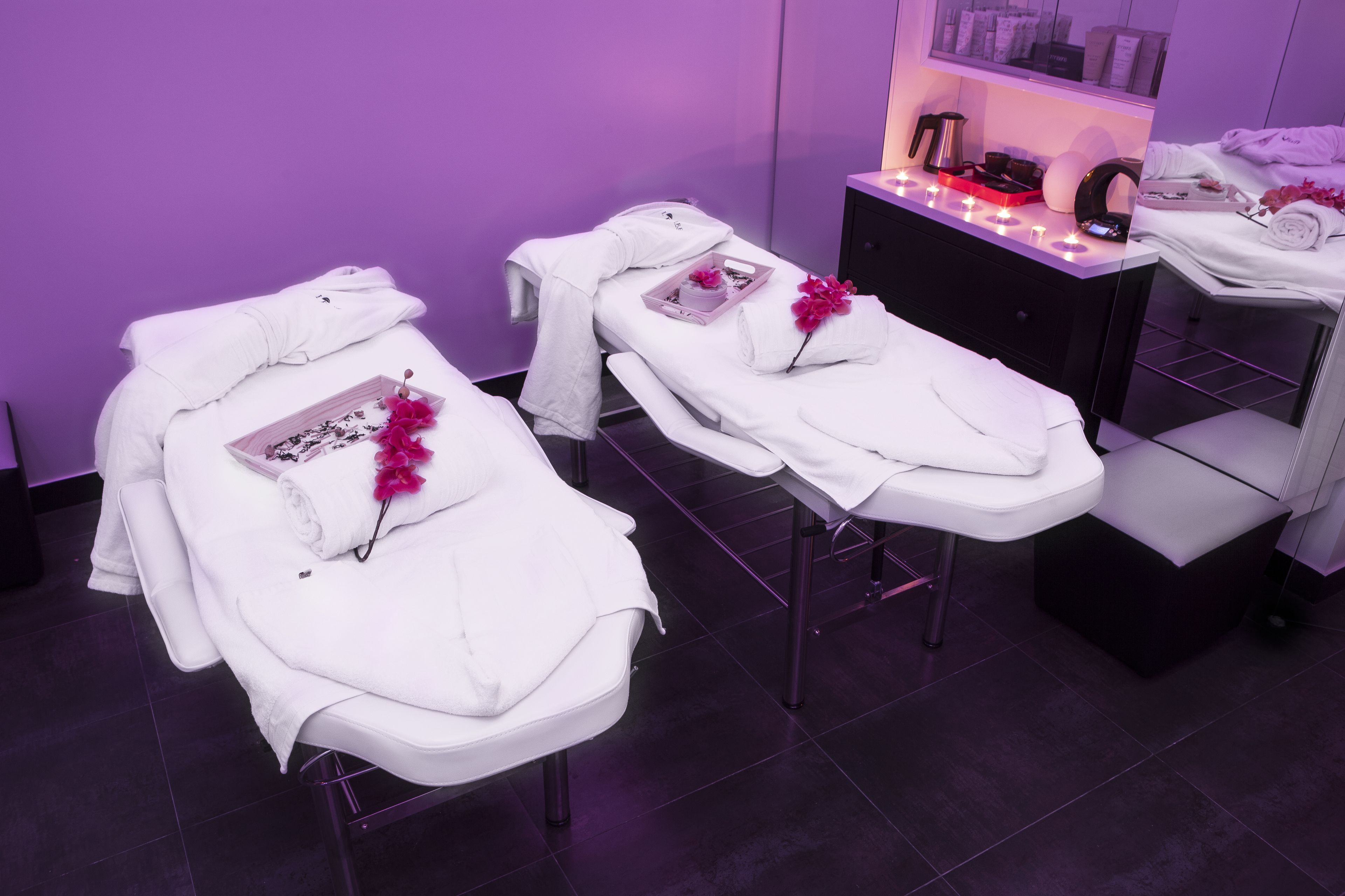 Un cuarto de spa con dos camillas, cada una con una toalla blanca y una flor rosa. Hay un mueble negro con una bandeja de objetos y una mesita con una vela.
