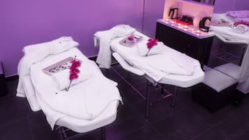 Ein Spa-Raum mit zwei Behandlungsbetten, jeweils mit einem weißen Handtuch und einer rosa Blume. Es gibt eine schwarze Schrank mit einer Ablage voller Gegenstände und einen kleinen Tisch mit einer Kerze.