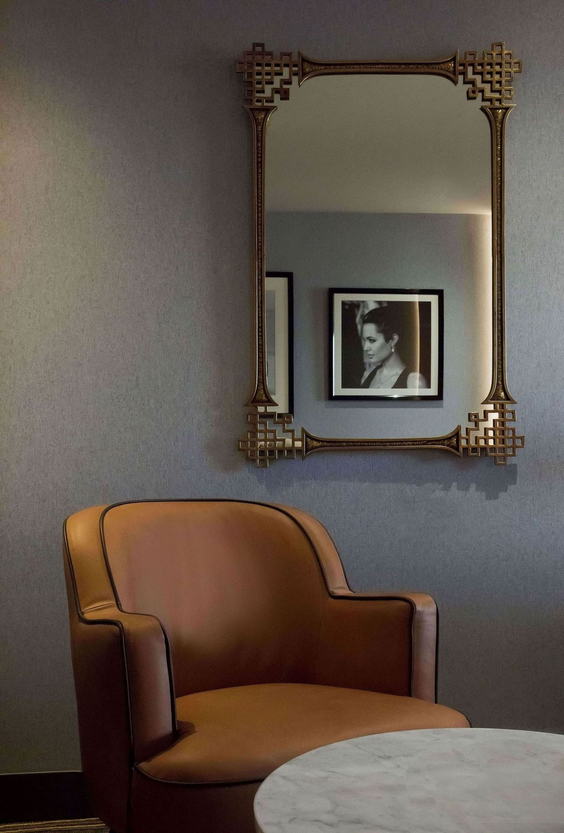 Una habitación con un espejo grande y ornamentado, una fotografía enmarcada en la pared, un sillón de cuero marrón y una mesa de mármol.