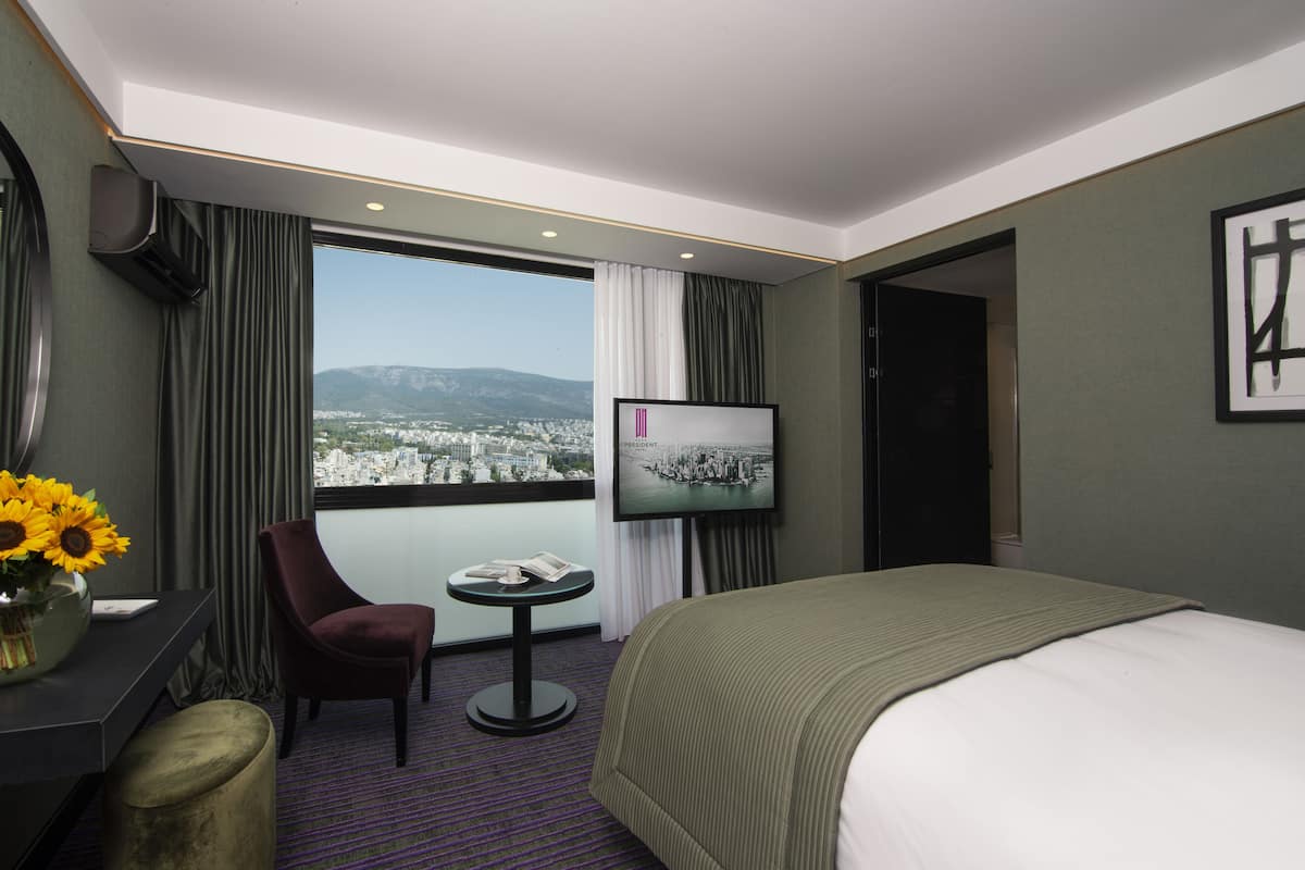 A hotel room with a large bed, a desk with a chair, a small table, a TV, and a view of a cityscape through a large window.