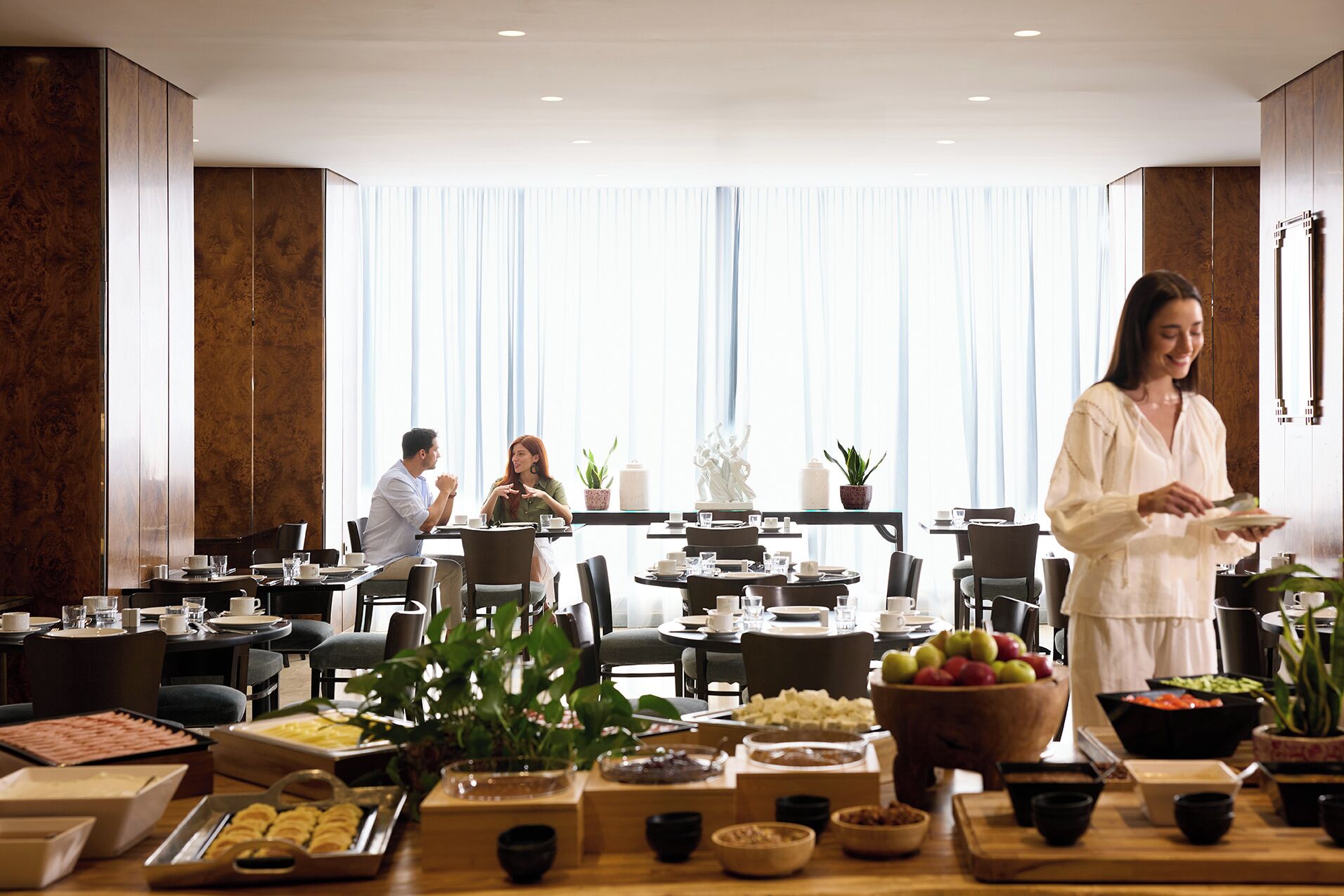A hotel dining area with multiple tables set for breakfast, a buffet with various food items, and a large window with curtains.