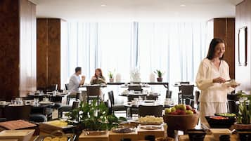 A hotel dining area with multiple tables set for breakfast, a buffet with various food items, and a large window with curtains.