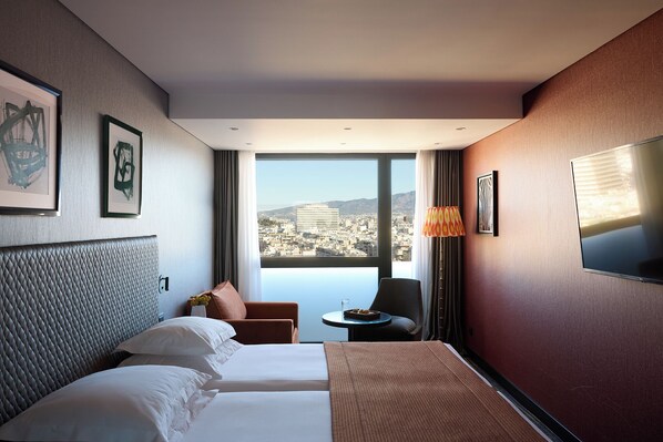 A hotel room with a bed, a chair, a small table, and a TV. There is a window with a city view.