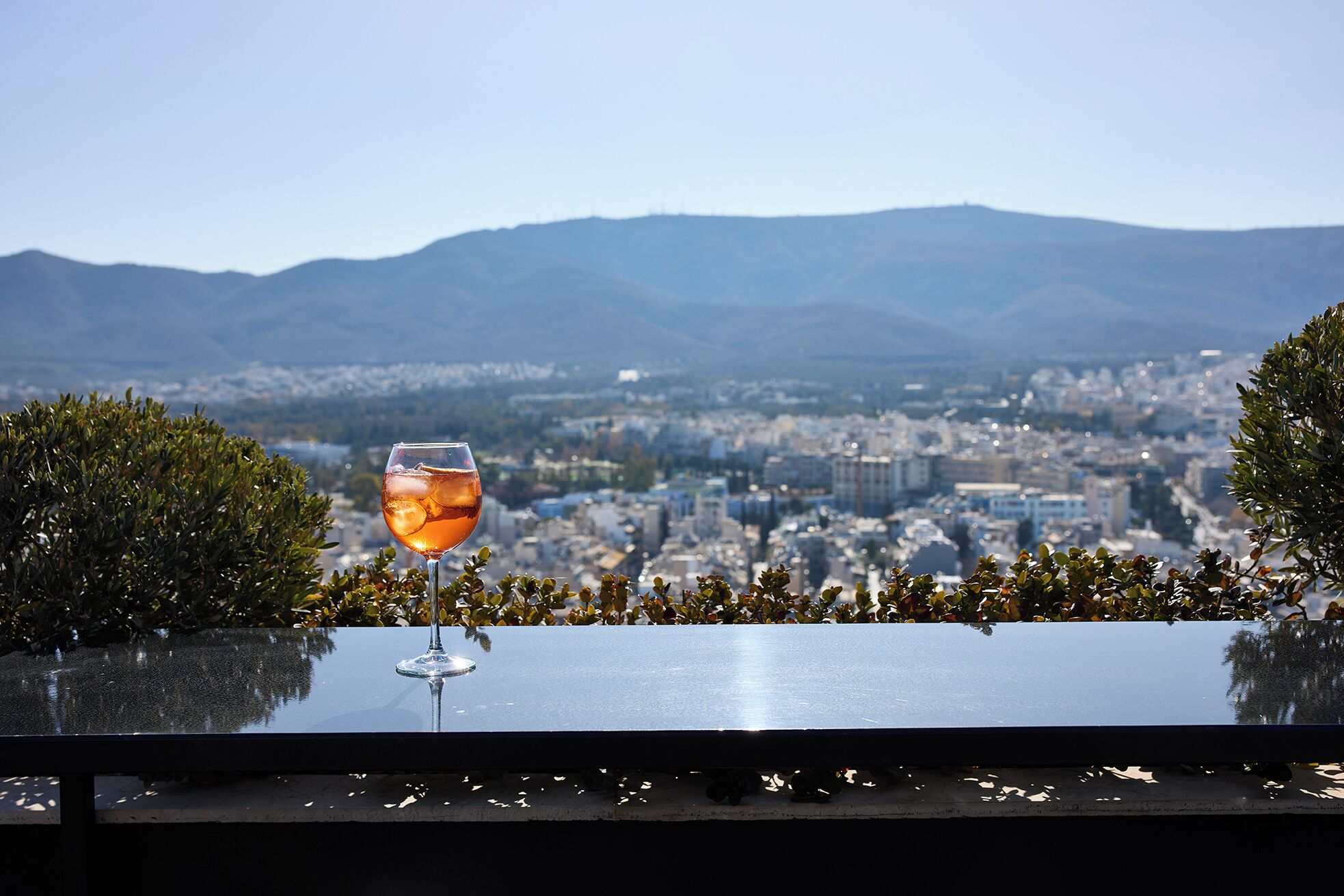 Un verre de vin sur un balcon donnant sur une vue panoramique de la ville, avec des montagnes en arrière-plan.