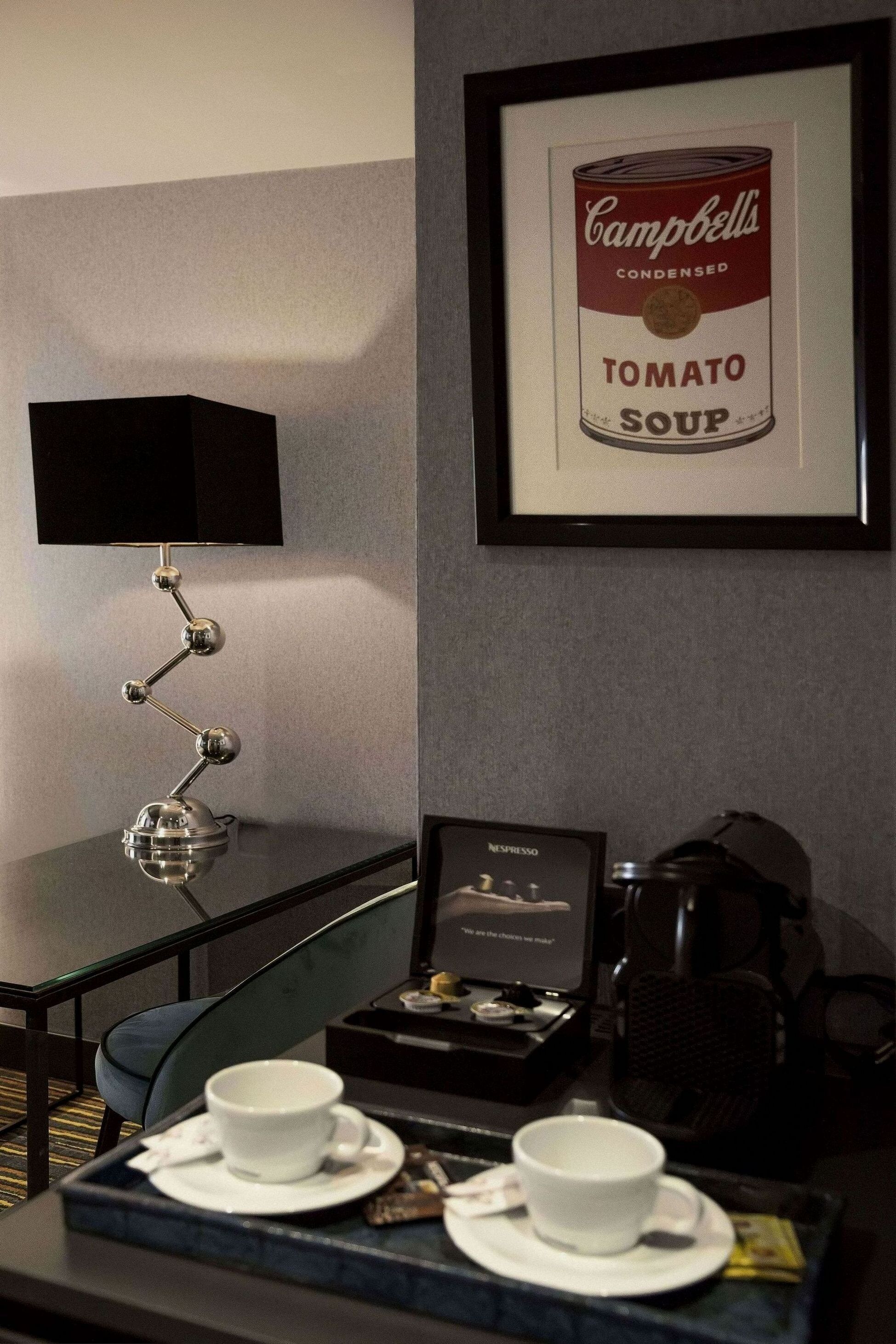 Una habitación de hotel con una lata enmarcada de sopa de tomate Campbell's, un escritorio con una lámpara, una máquina de café y una bandeja con tazas y una caja de cápsulas Nespresso.