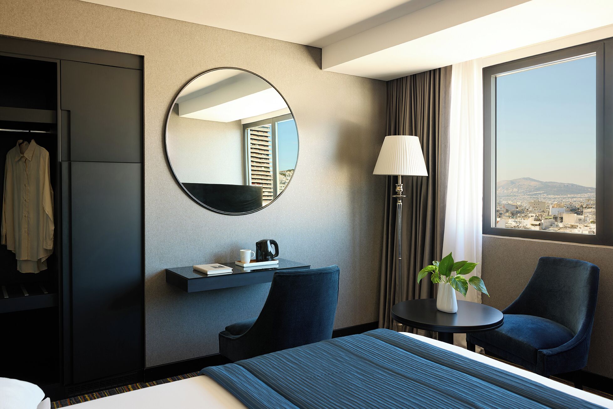 Une chambre d’hôtel avec un grand lit, un bureau avec une lampe, une chaise, un miroir et une vue sur le paysage urbain.