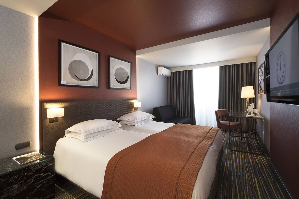 A hotel room with a large bed, two framed artworks on the wall, a desk with a lamp, and a TV.
