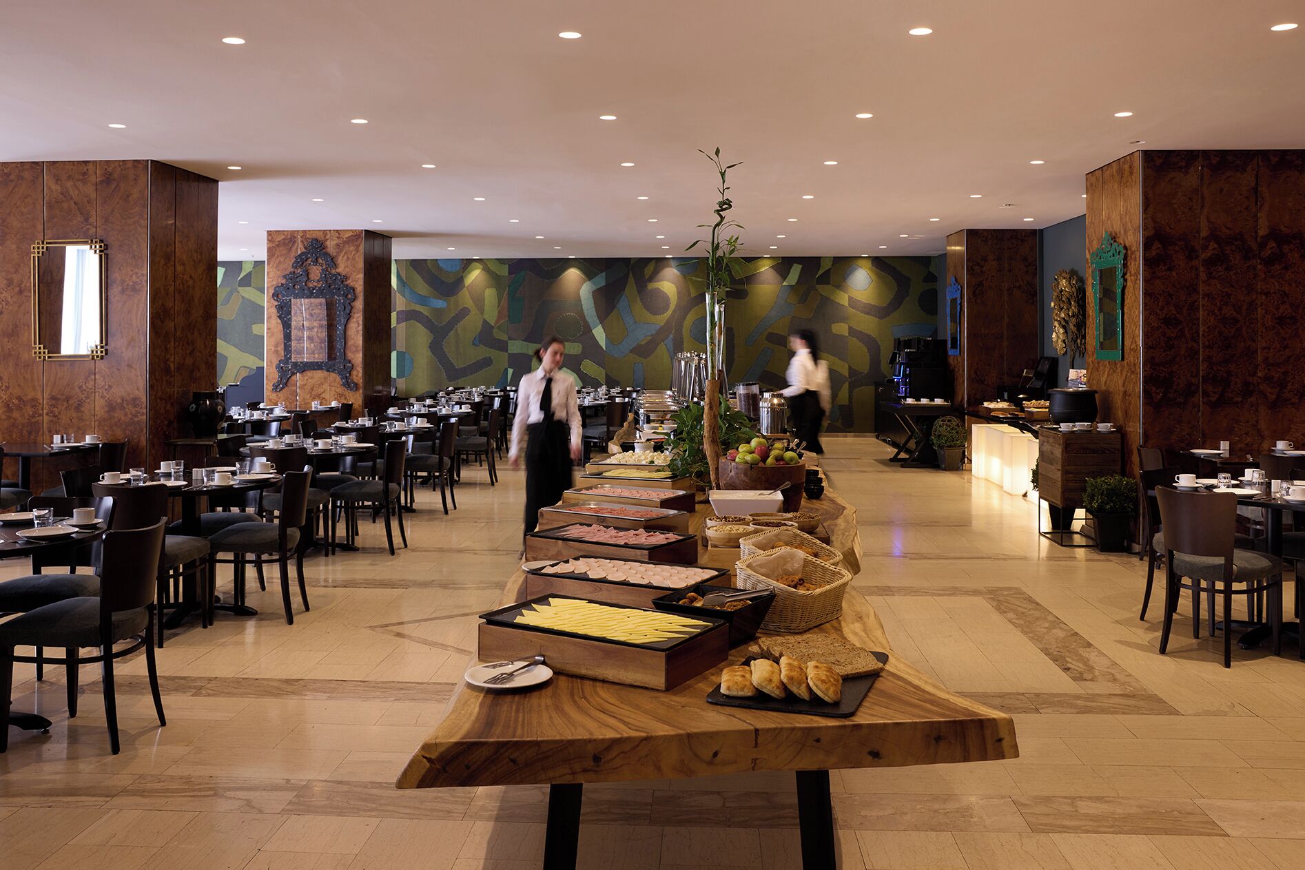 A buffet setup in a hotel dining area with multiple tables and chairs, a central buffet table with various food items, and a staff member in the background.