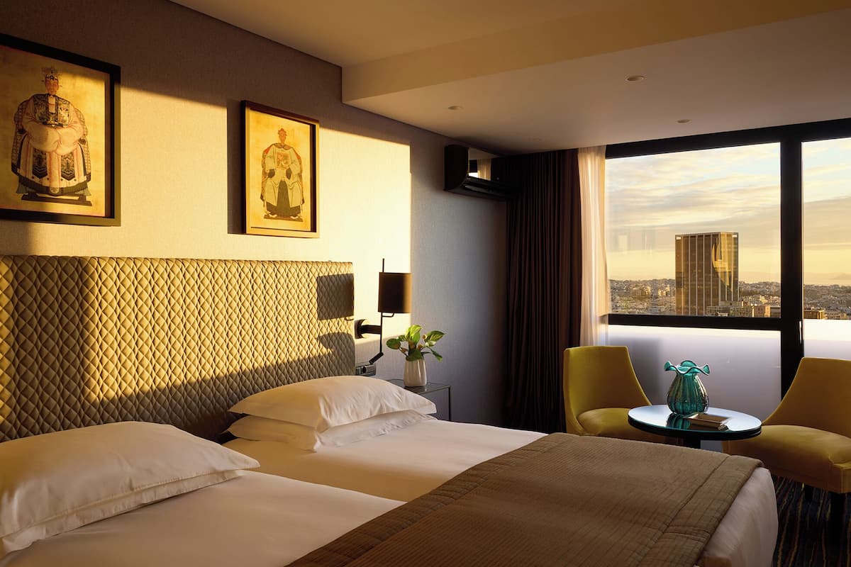 A hotel room with two beds, a view of a cityscape, and a decorative vase on a small table.