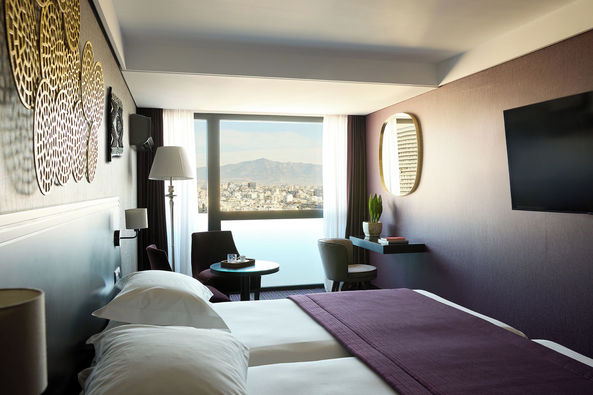A hotel room with a bed, a desk, a chair, a small table, a lamp, a mirror, and a view of a cityscape.
