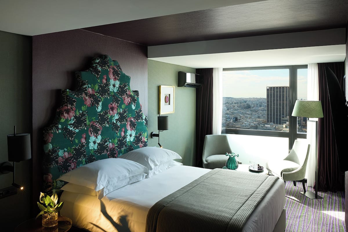 A hotel room with a large bed, a view of the city, a chair, and a lamp.