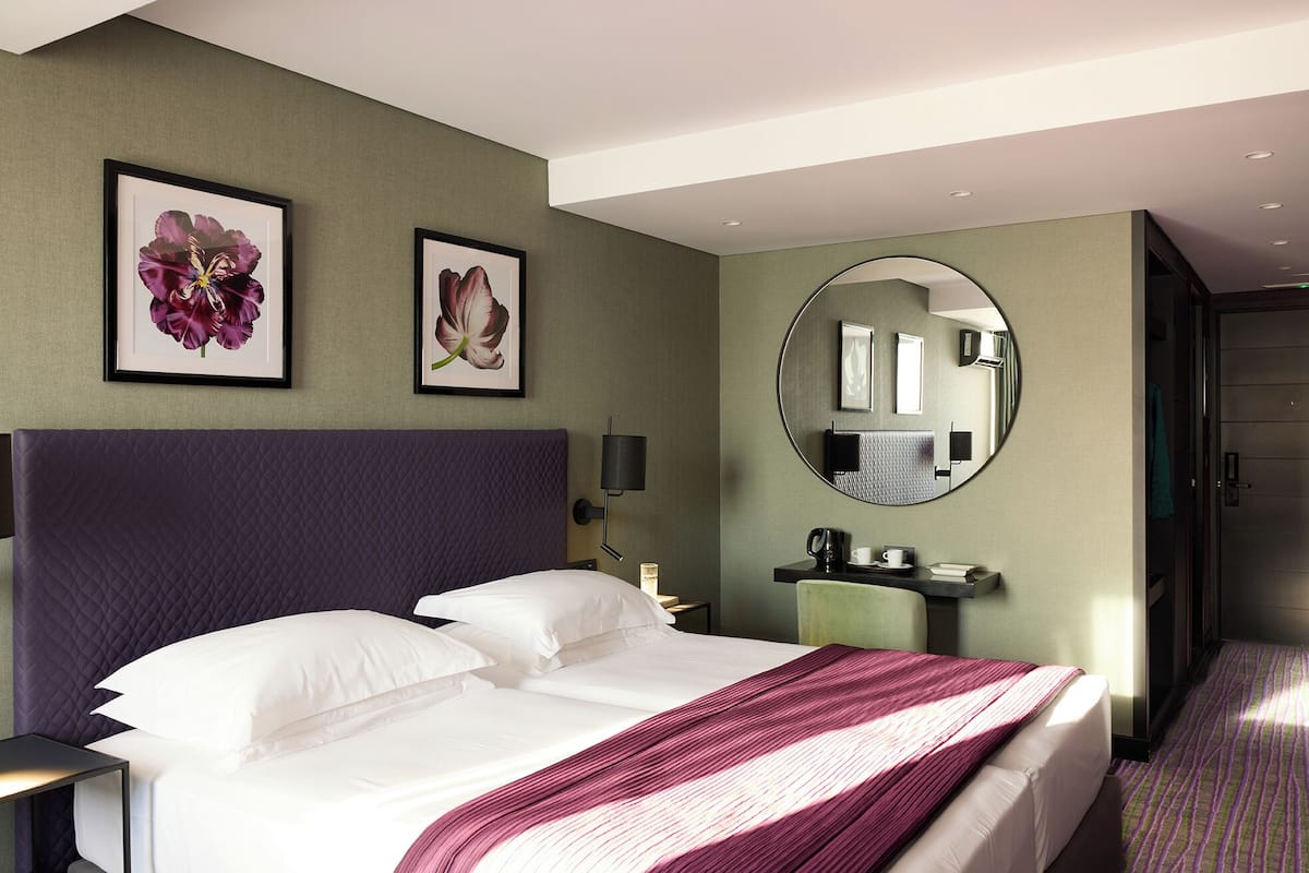 A hotel room with a large bed, a round mirror, two framed floral pictures, and a nightstand with a lamp.