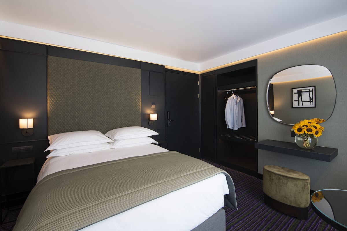 A hotel room with a large bed, a nightstand, a wardrobe, a mirror, and a small stool.