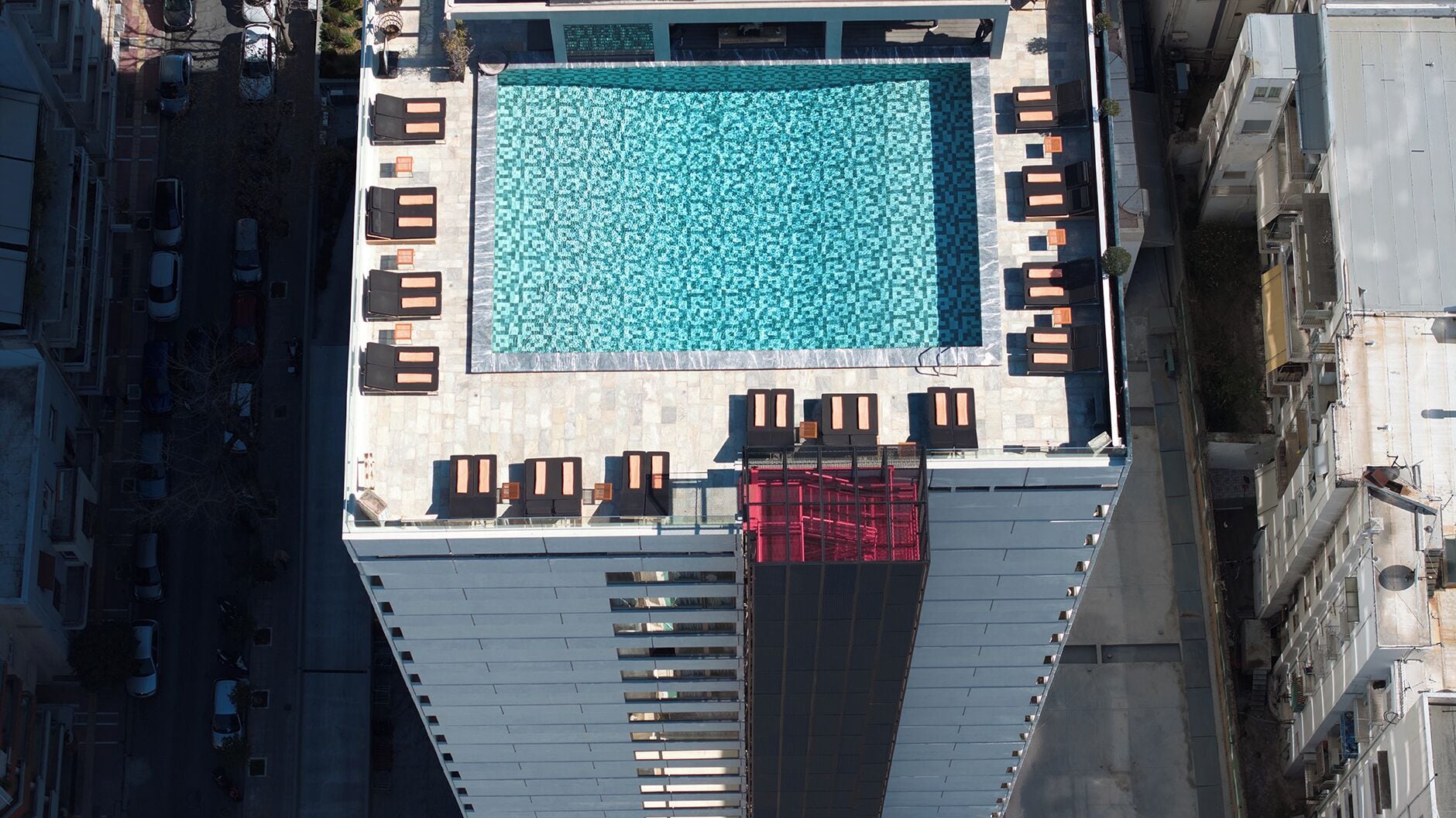 An aerial view of a rooftop pool with a geometric tile pattern, surrounded by sun loungers and a covered seating area.