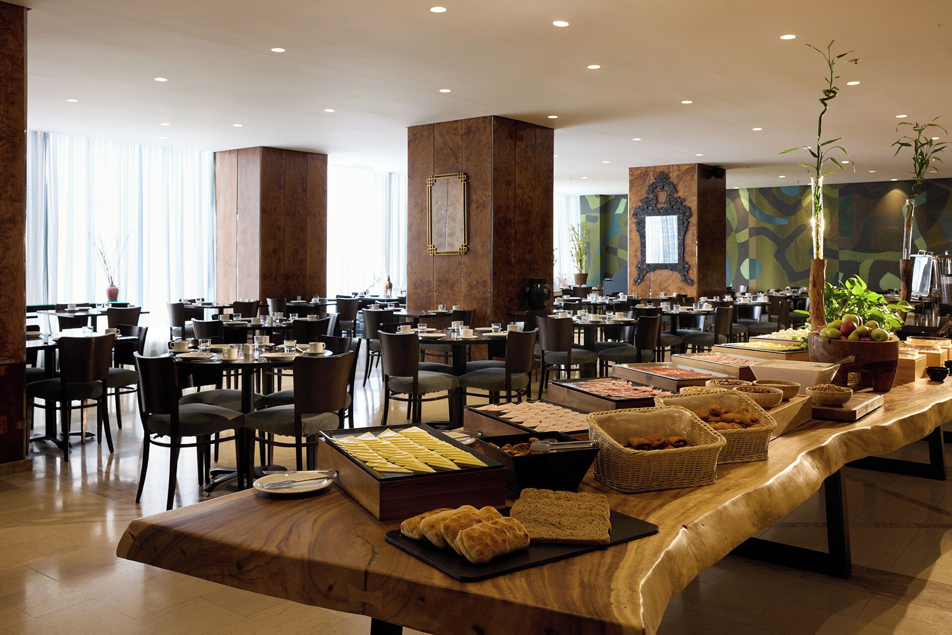 A spacious dining area with multiple tables and chairs, a buffet with various food items, and a large wooden table with bread and pastries.