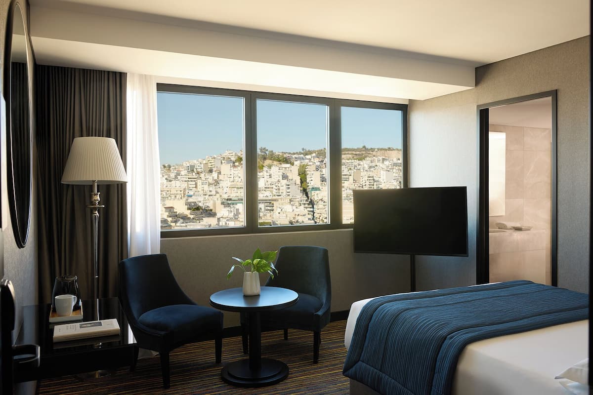 A hotel room with a bed, a TV, a desk with a chair, a lamp, and a view of a cityscape.
