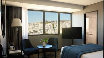 Habitación de hotel con cama, televisor, escritorio con silla, lámpara y vista a un paisaje urbano.