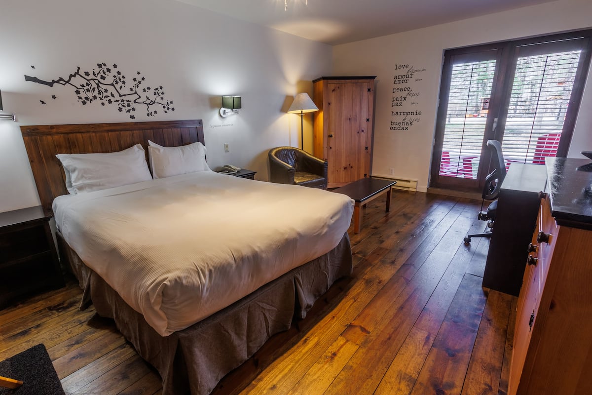 A hotel room with a large bed, a wooden headboard, a desk with a chair, a wardrobe, a lamp, and a wall with inspirational quotes.