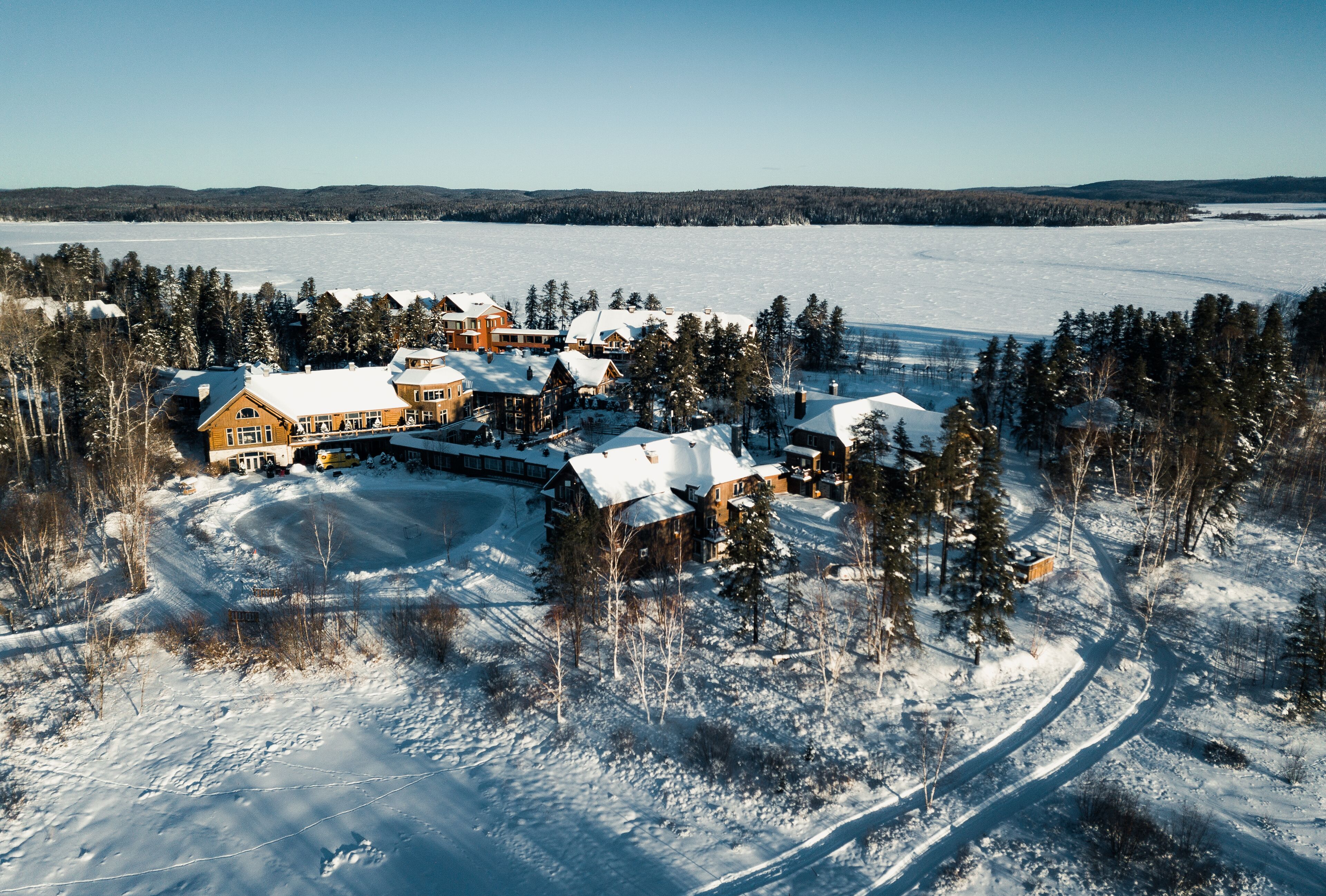 Un gran lodge con varias construcciones rodeado de árboles cubiertos de nieve y un lago congelado al fondo.