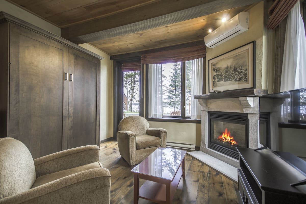 A cozy living room with a fireplace, wooden furniture, and large windows offering a view of trees.