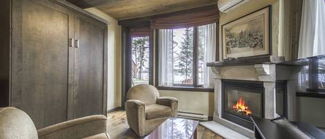 Un salón acogedor con chimenea, muebles de madera y ventanas grandes que dan a un paisaje de árboles.