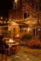 Un edificio de madera con fachada de piedra, mesas al aire libre y luces colgantes.
