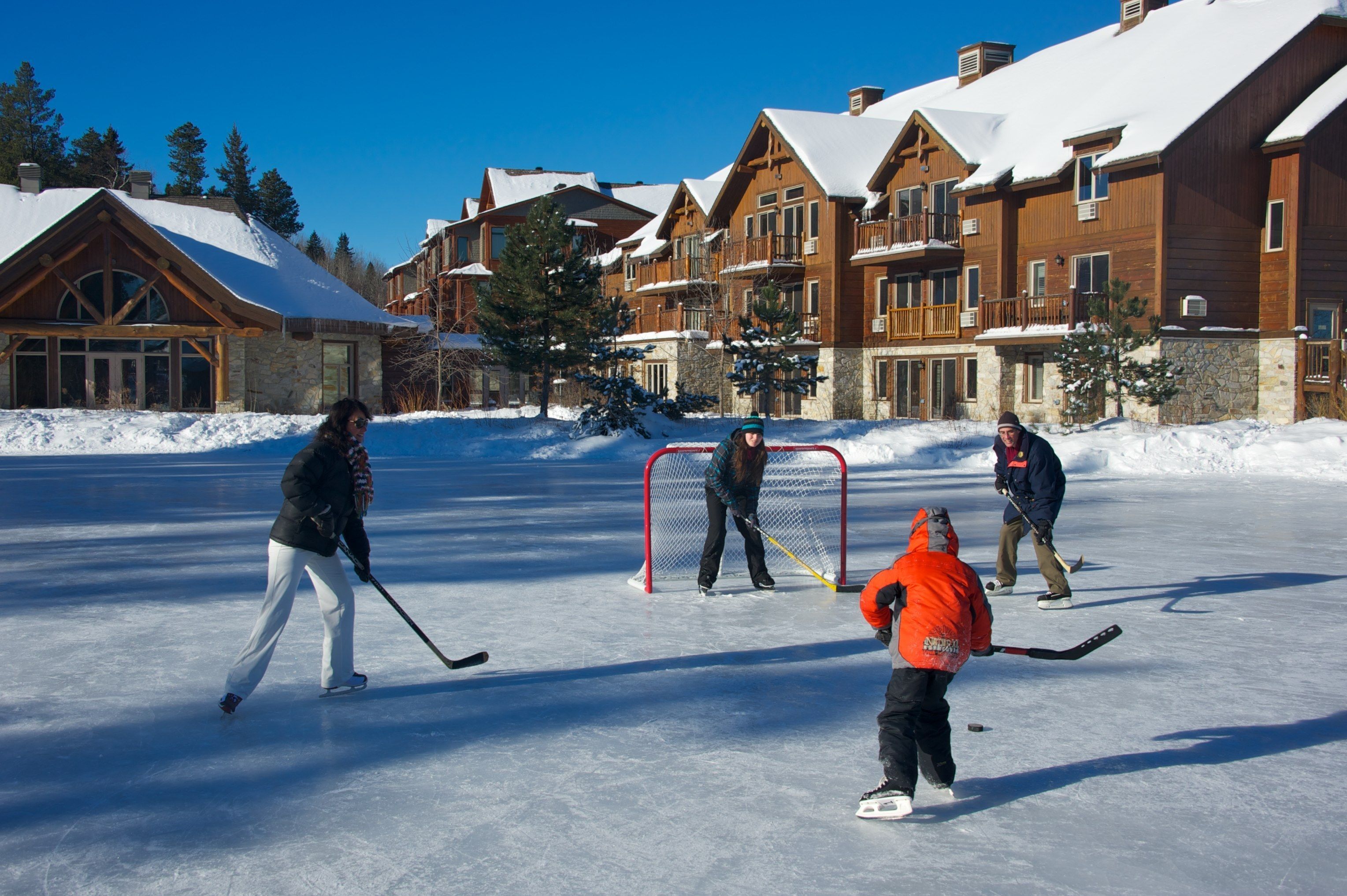 A group of people ice skating and playing hockey on a frozen surface with wooden buildings in the background.