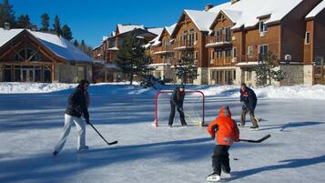 A group of people ice skating and playing hockey on a frozen surface with wooden buildings in the background.