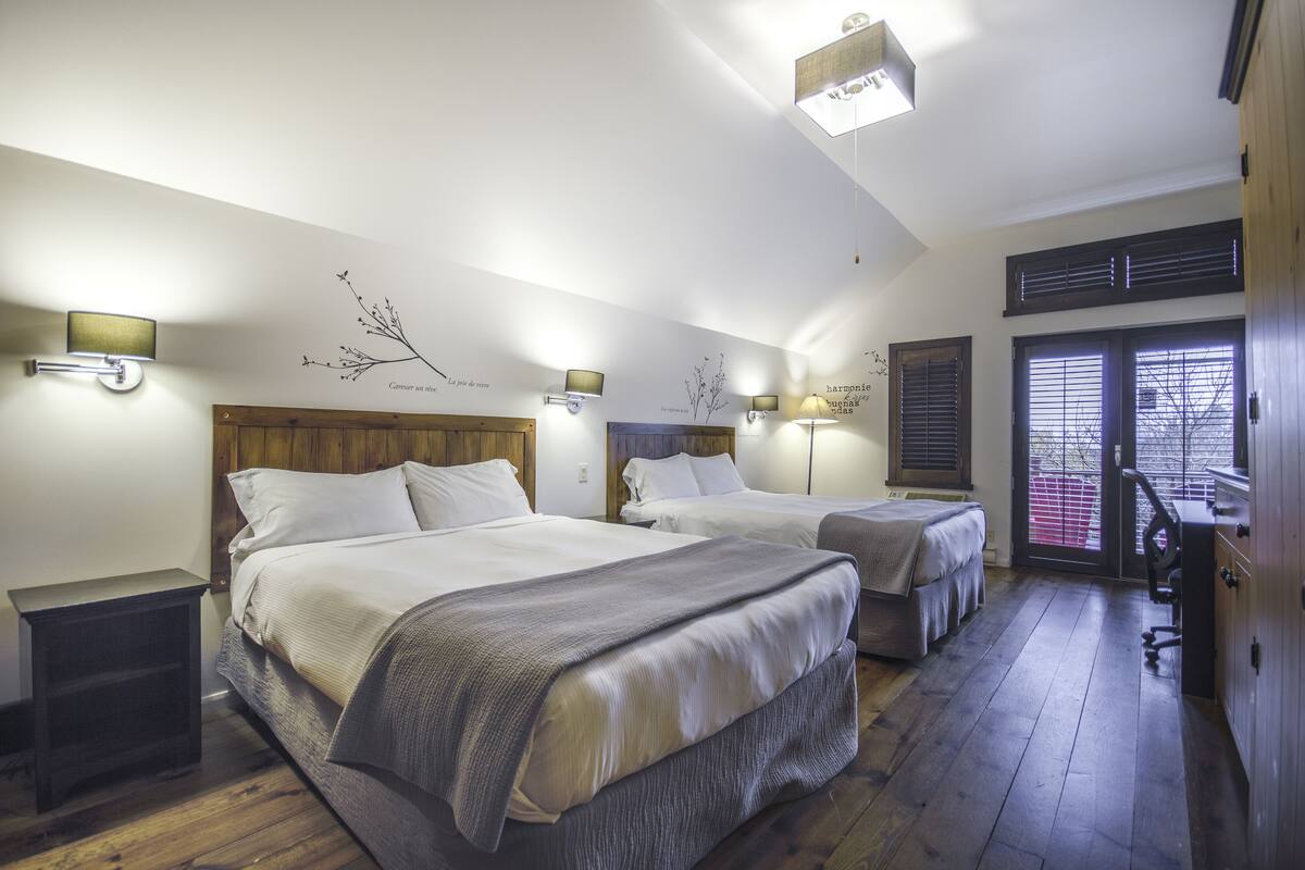 A hotel room with two beds, wooden flooring, and wall-mounted lamps.
