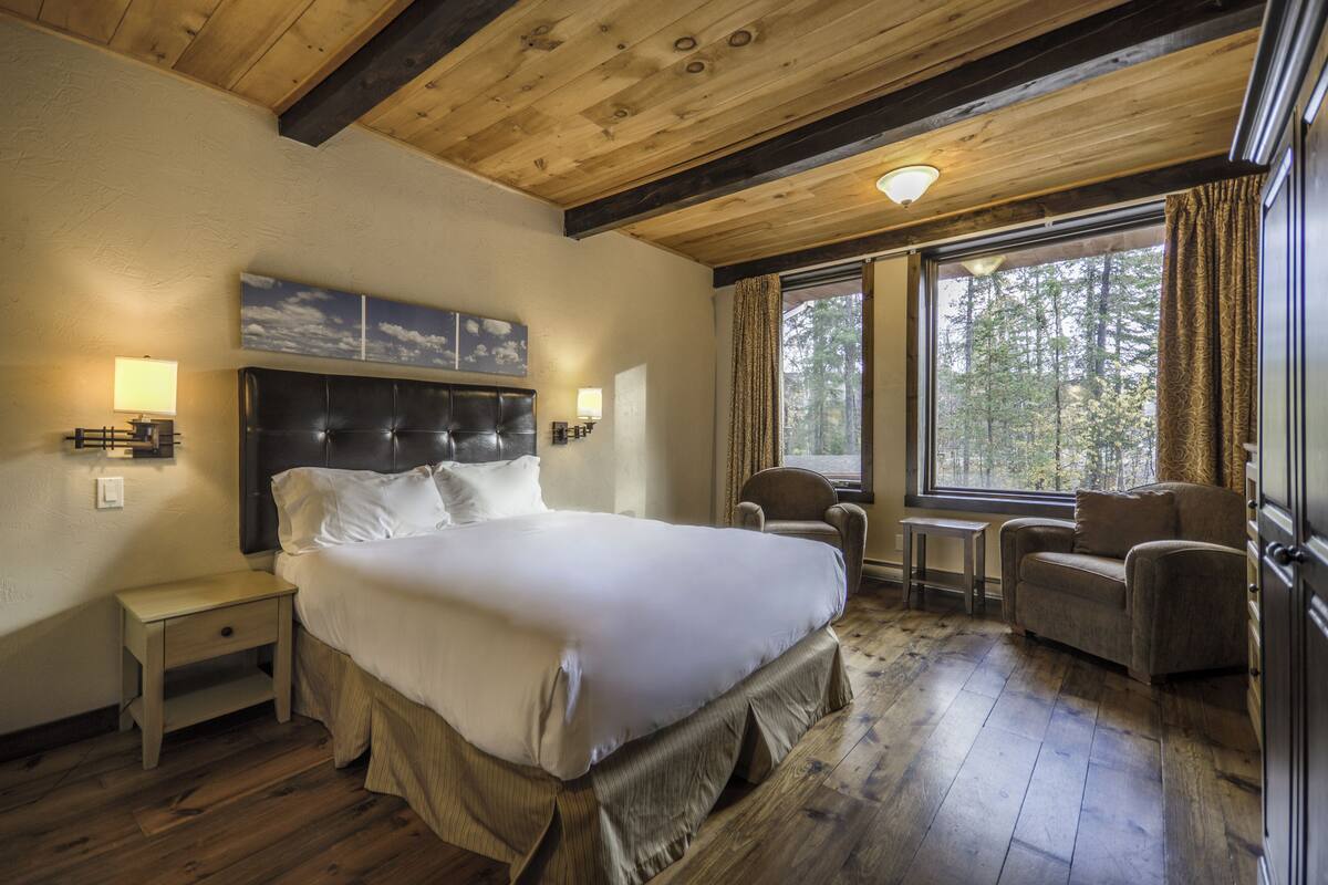 A bedroom with a large bed, a wooden ceiling, a window with curtains, a chair, and a small table.
