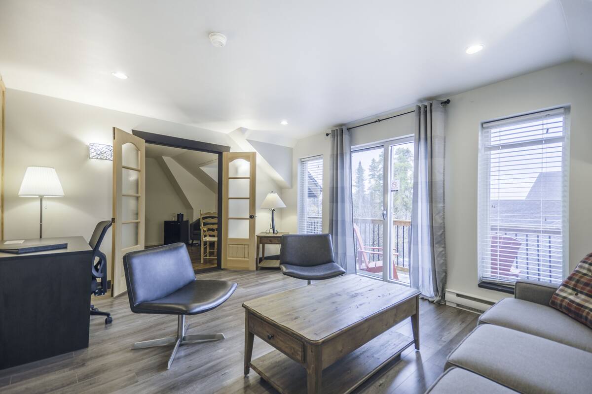 A modern living room with a wooden coffee table, two black chairs, a sofa, a desk with a lamp, and a staircase leading to another level.