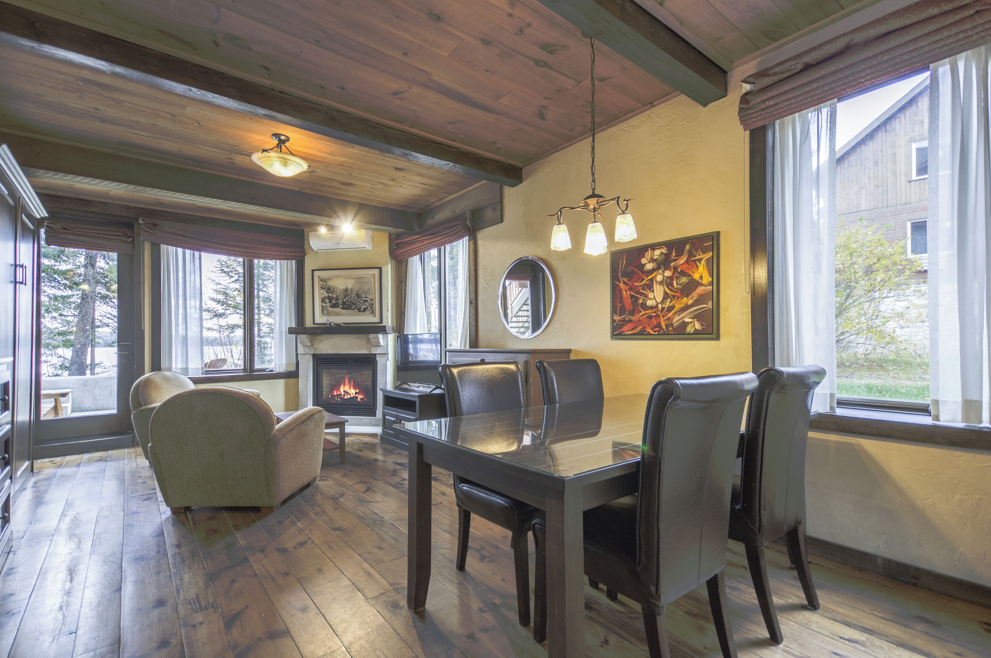 A cozy living room with a fireplace, wooden floors, a dining table with chairs, and large windows offering a view of the outside.