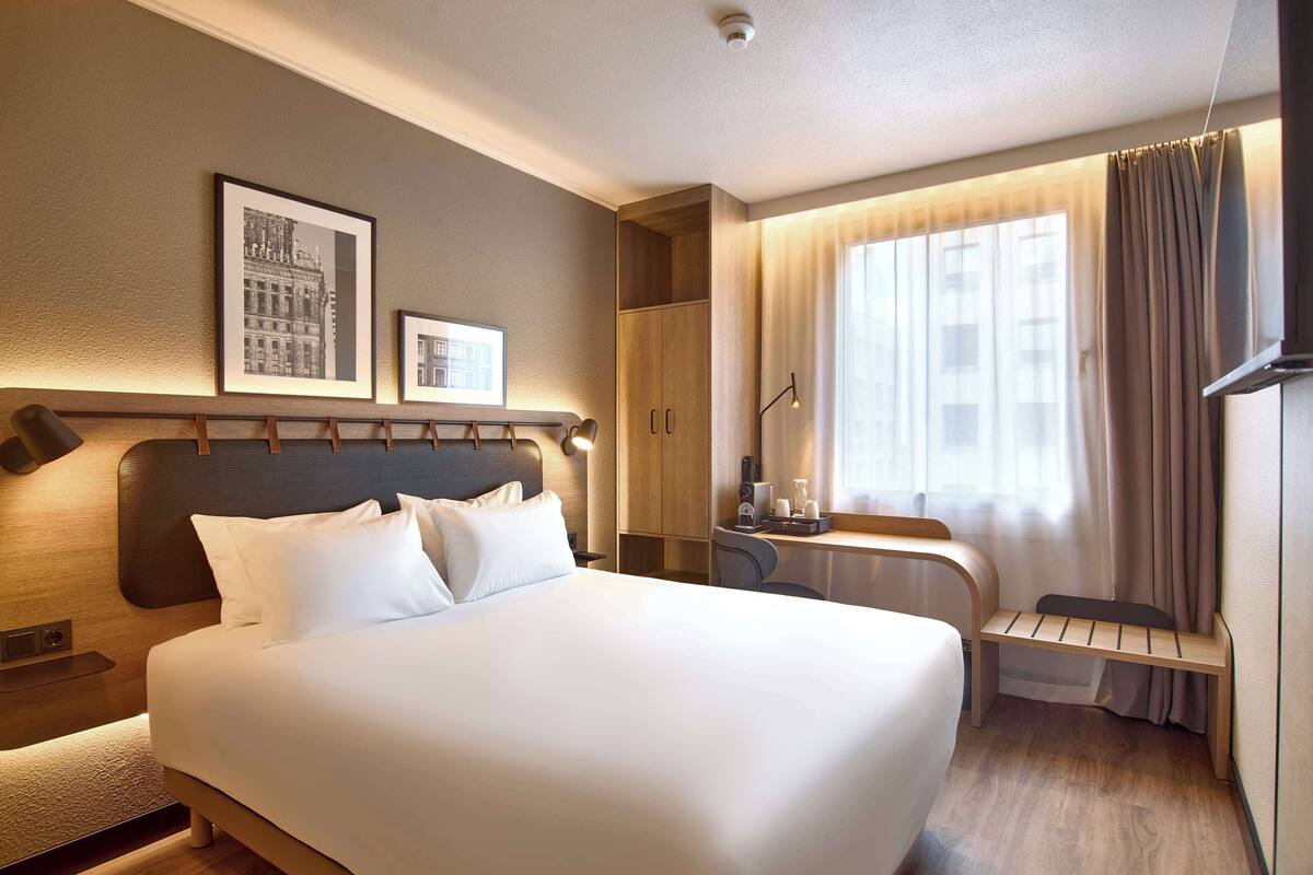 Une chambre d’hôtel avec un grand lit, un bureau avec une chaise, une armoire et deux tableaux encadrés au mur.