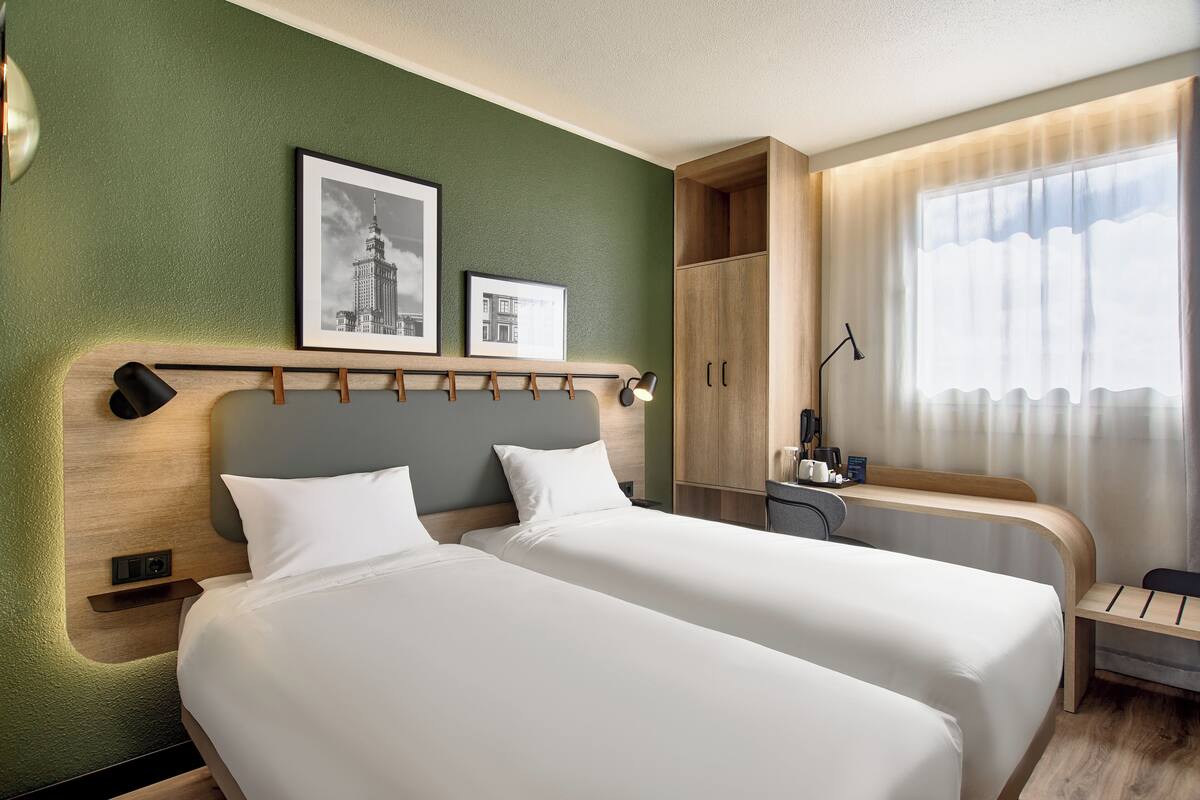 Une chambre d’hôtel avec deux lits, un bureau, une chaise et une fenêtre avec des rideaux légers.