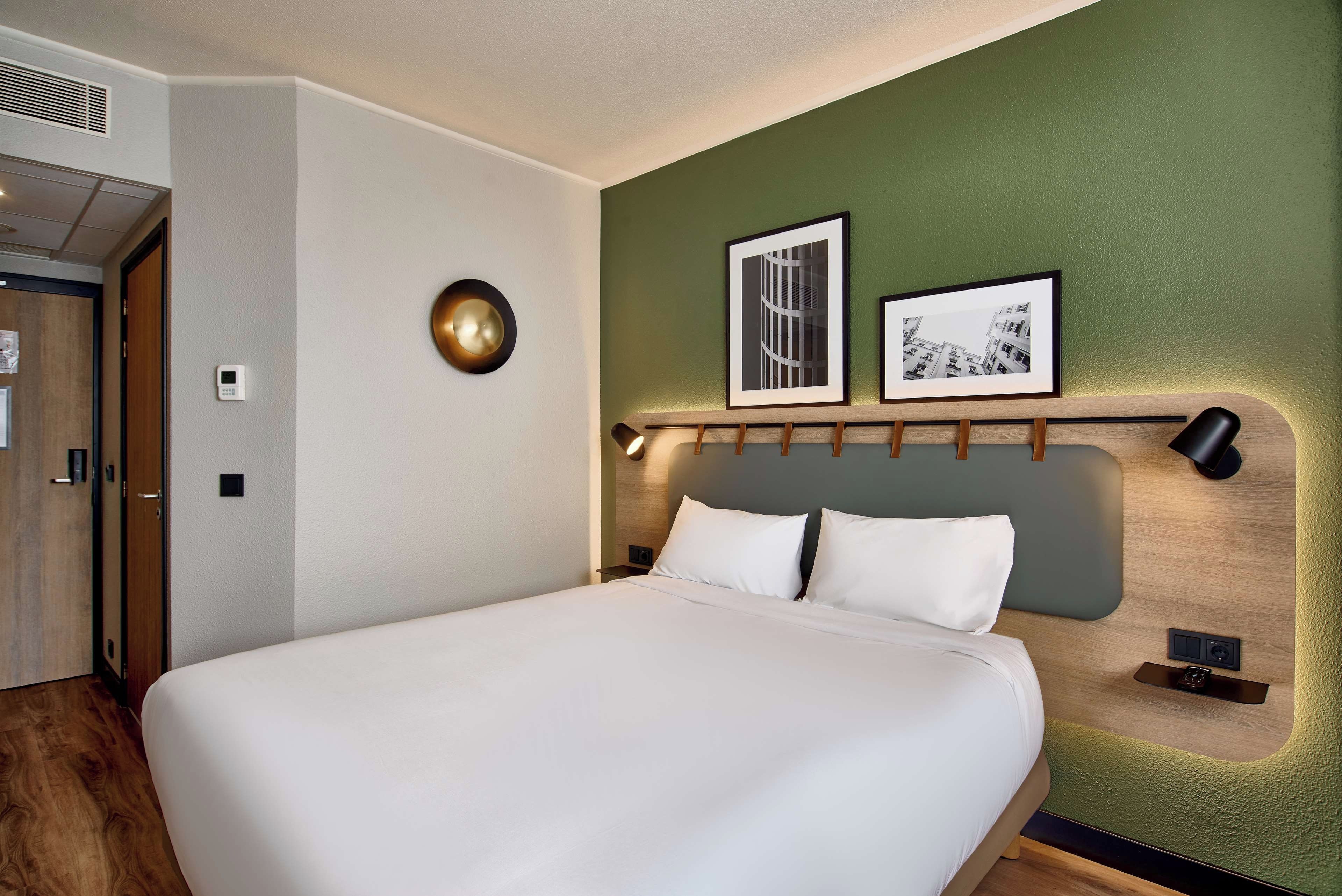 Habitación de hotel con cama, cabecera con iluminación integrada, rejilla de ventilación en la pared y dos cuadros enmarcados.