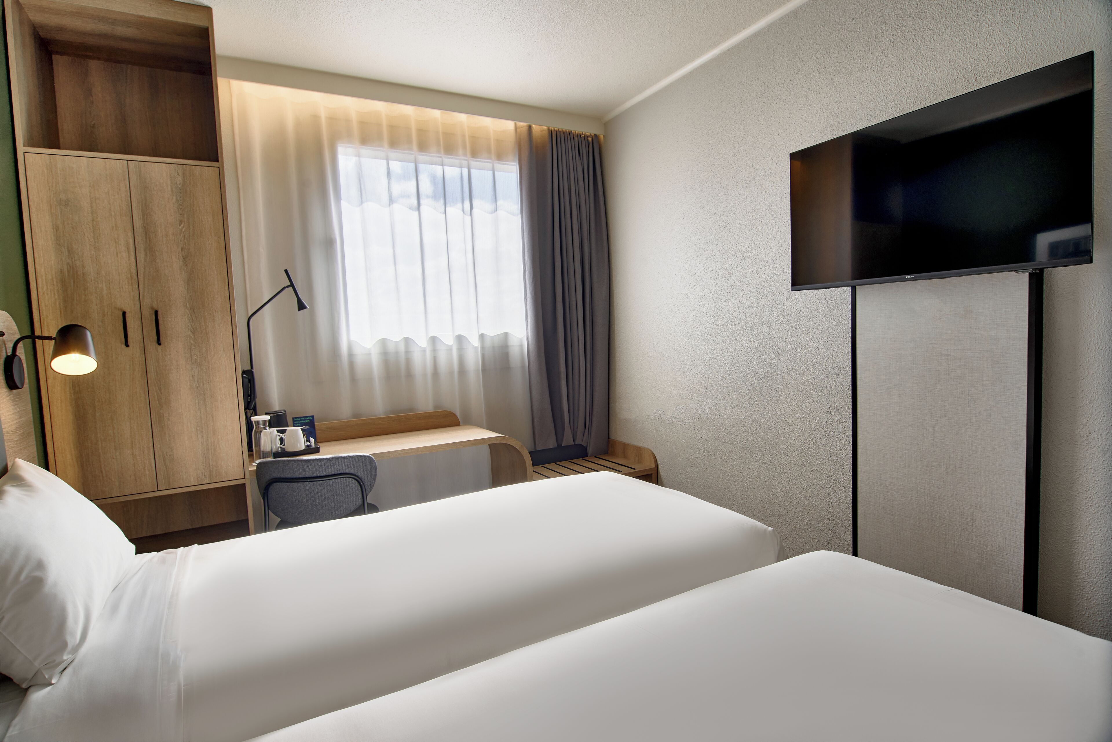 A hotel room with a bed, a desk with a chair, a TV, and a wardrobe.