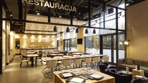 Un restaurante moderno con techo alto, paredes de ladrillo visto y grandes ventanales. Mesas y sillas dispuestas para comer.
