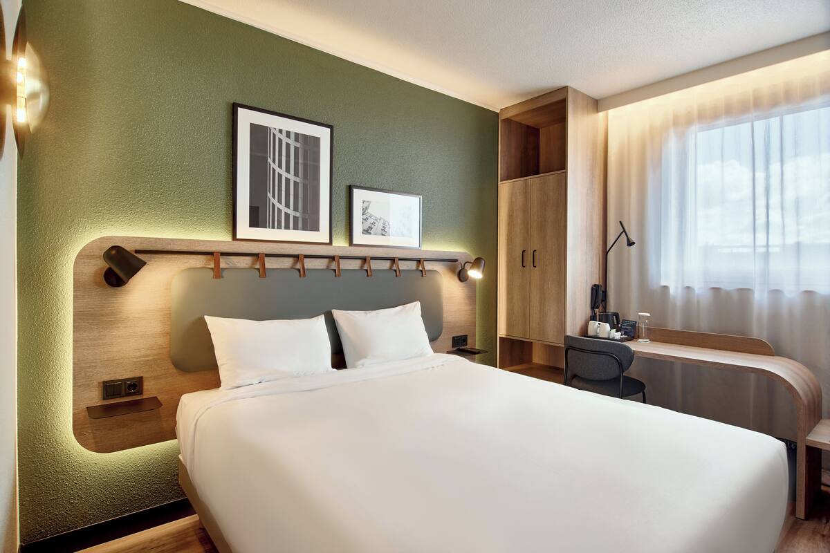 Une chambre d’hôtel avec un grand lit, un bureau avec une chaise, une armoire et des tableaux encadrés au mur.