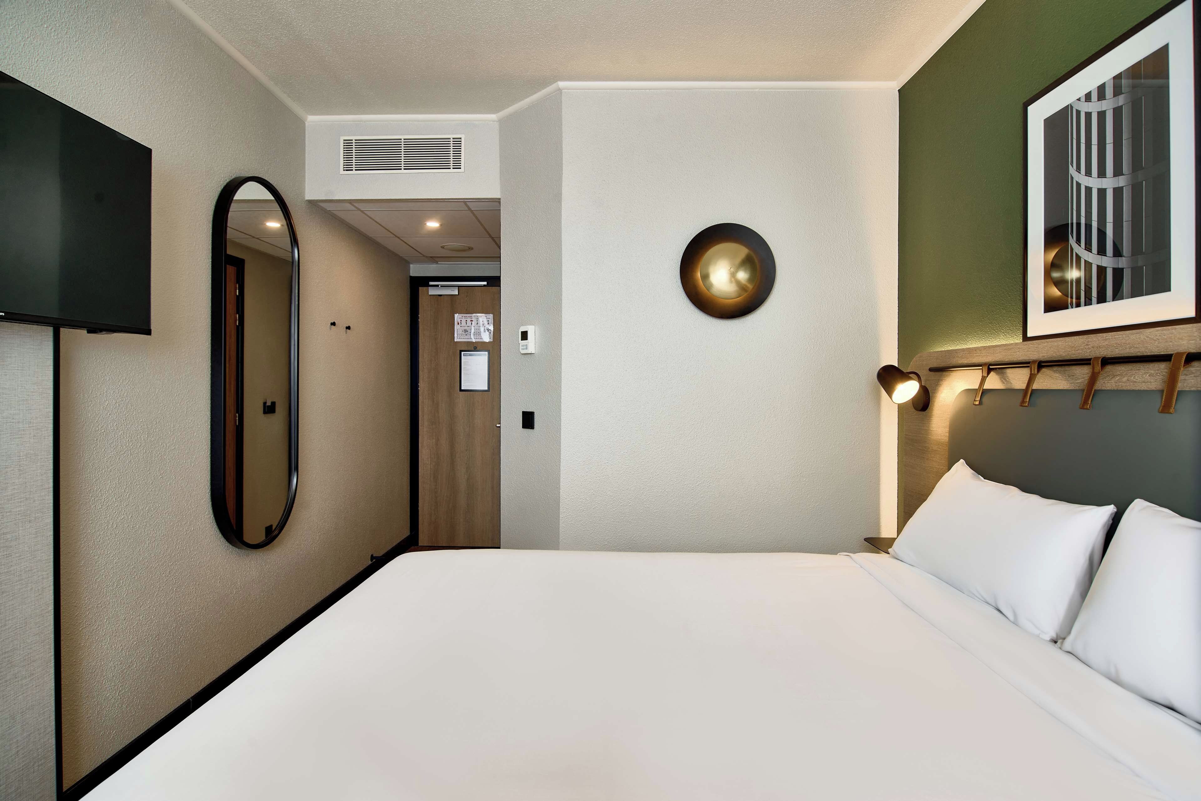 Une chambre d’hôtel comprenant un lit, une télévision, un miroir, un luminaire fixé au mur et une porte donnant sur une autre pièce.