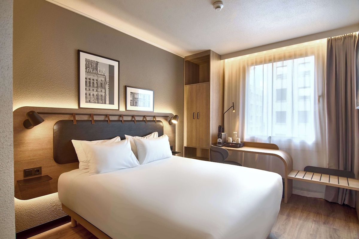Habitación de hotel con una cama grande, un escritorio con silla, un armario y dos cuadros en la pared.