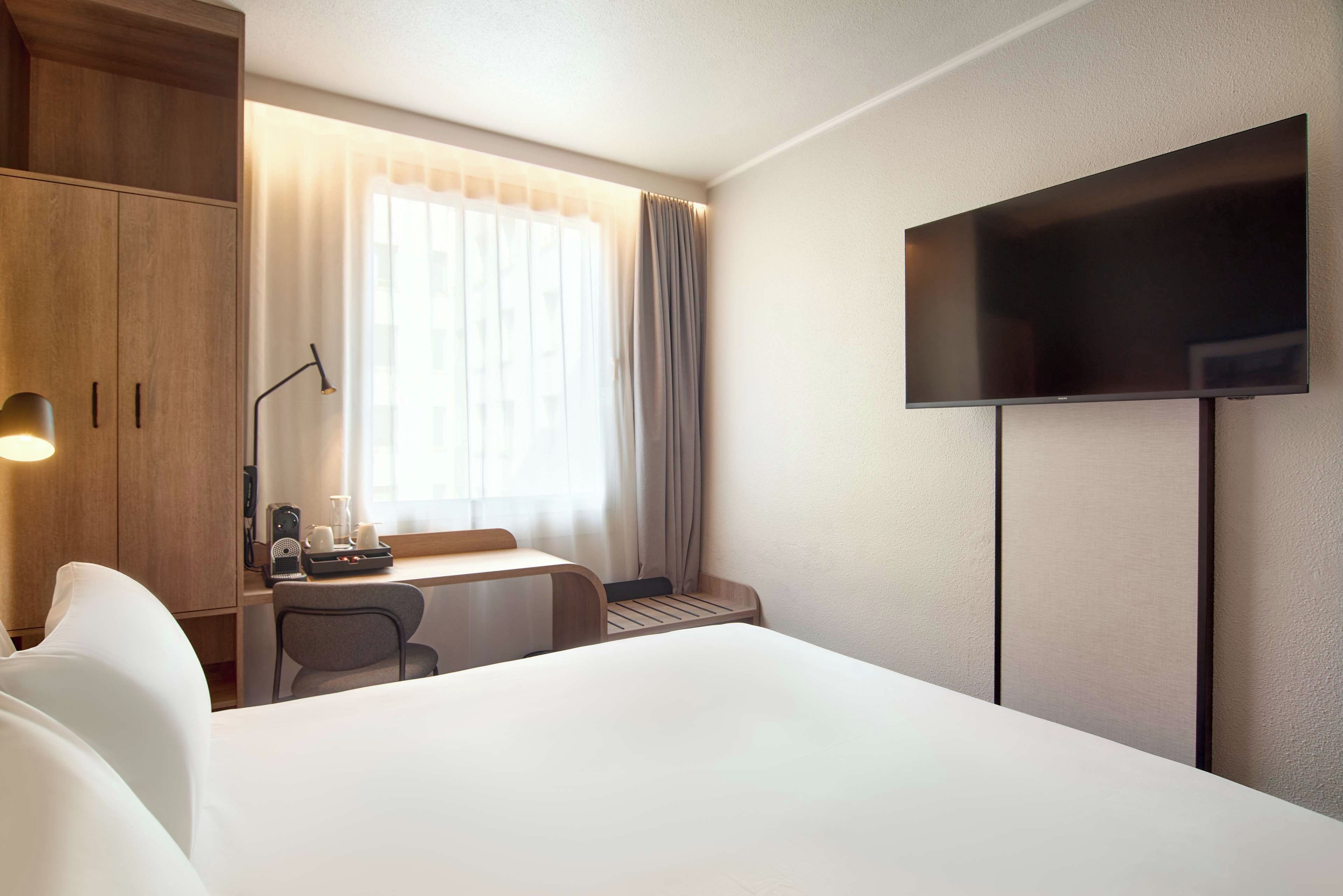 A hotel room with a bed, a desk with a chair, a television, and a window with curtains.