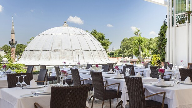 Un área de comedor al aire libre con mesas dispuestas para una comida, rodeada de vegetación y una gran estructura en forma de cúpula blanca al fondo.