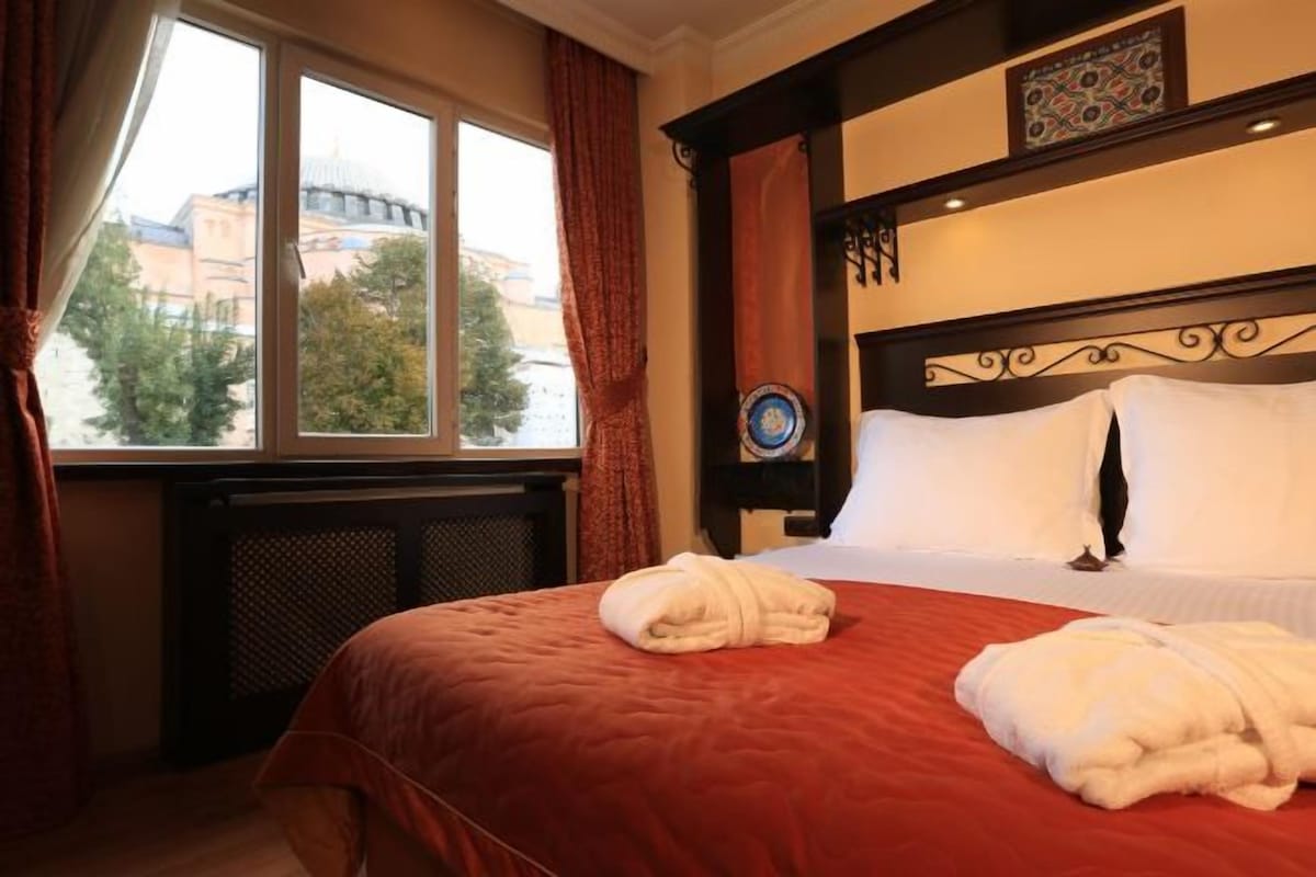 A hotel room with a large bed, a window with curtains, and a view of buildings outside.